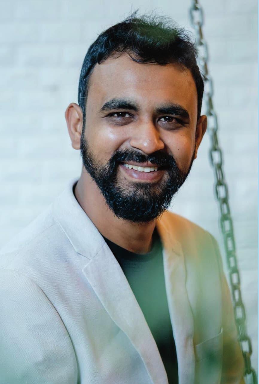 Gaurav Shewale, smiling, in a light jacket against a white brick wall